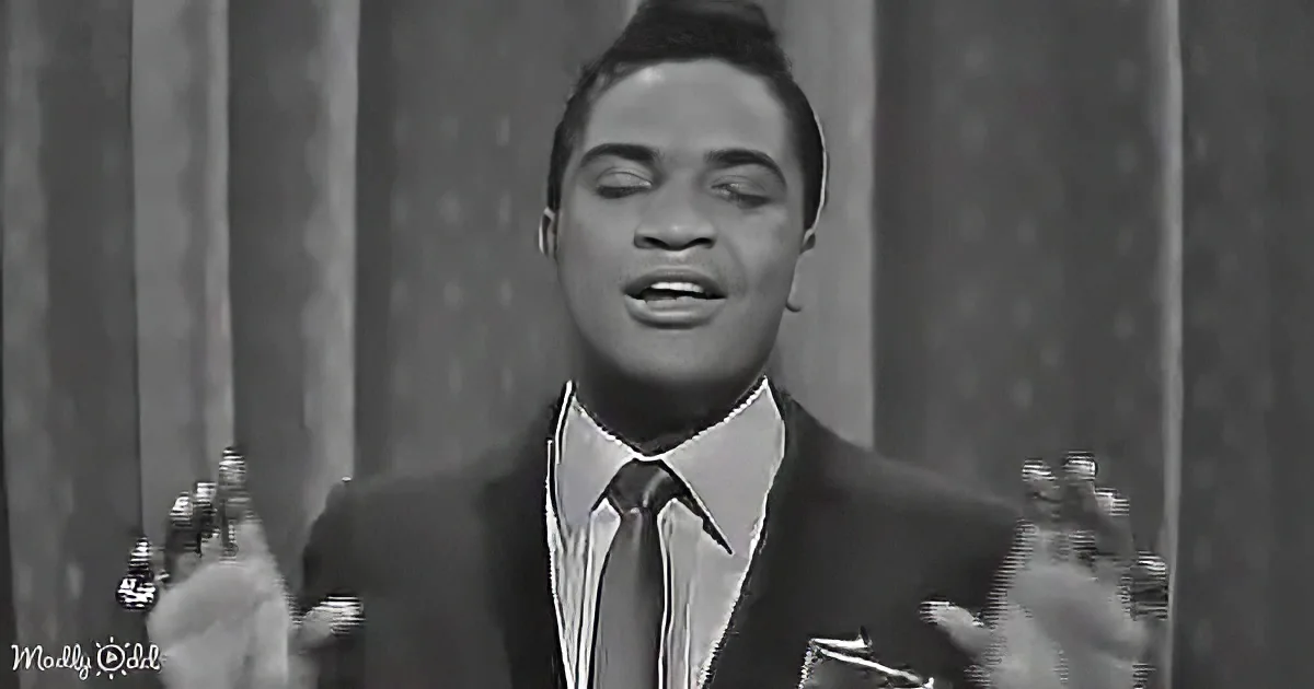 A man in a suit and tie is singing, hands raised, in front of a curtain backdrop.