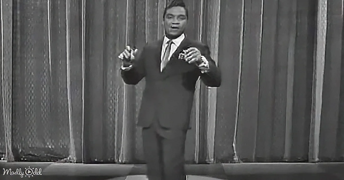 A man in a suit and tie stands on stage with his hands raised, possibly singing or performing.