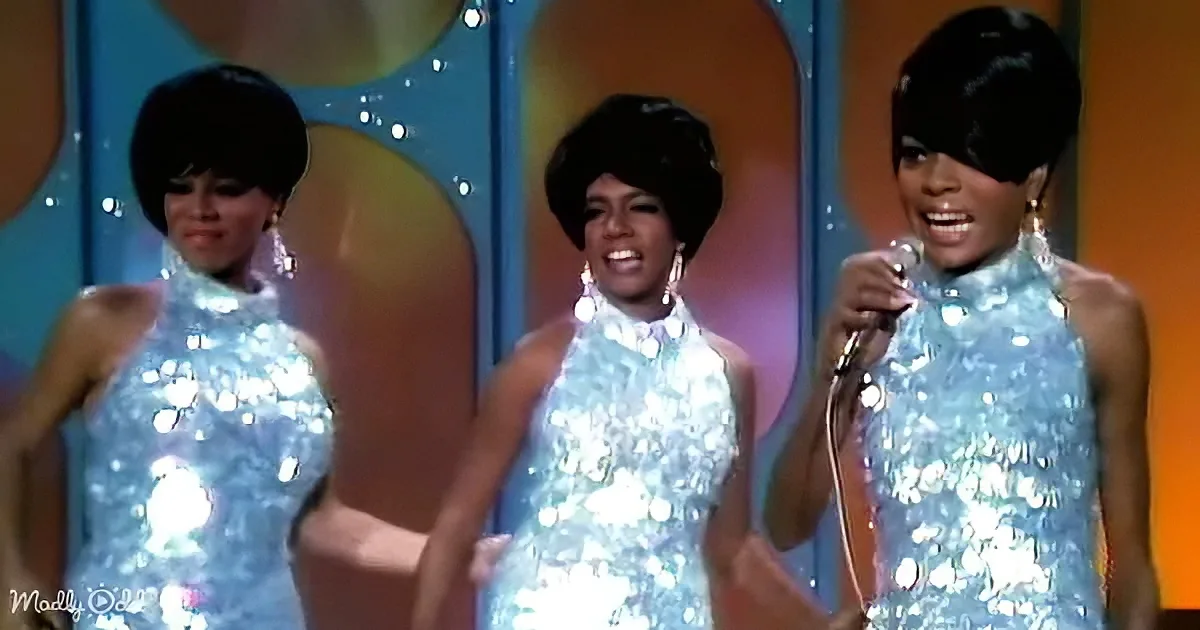 Three women in shiny blue dresses perform on stage with a vibrant, colorful background.