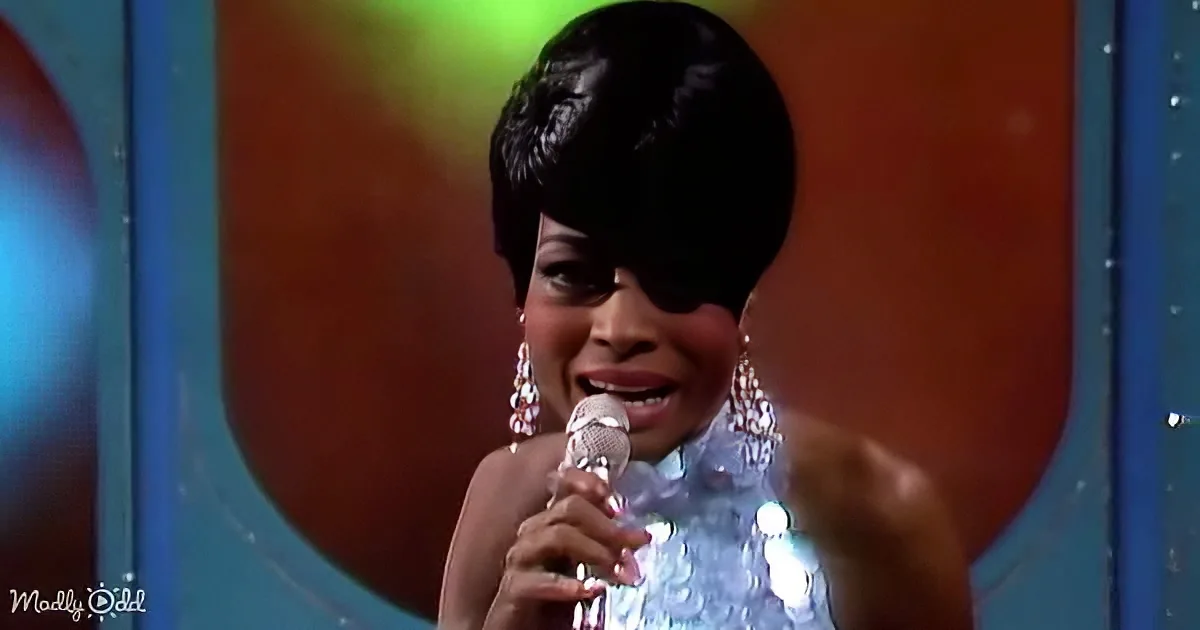 A woman with a short black hairstyle and large earrings sings into a microphone, wearing a sparkly outfit.