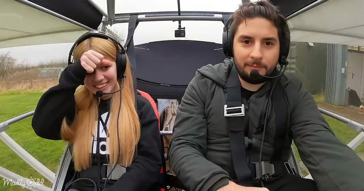 Two individuals wearing headsets are seated in a small aircraft, with a grassy field and building visible outside.