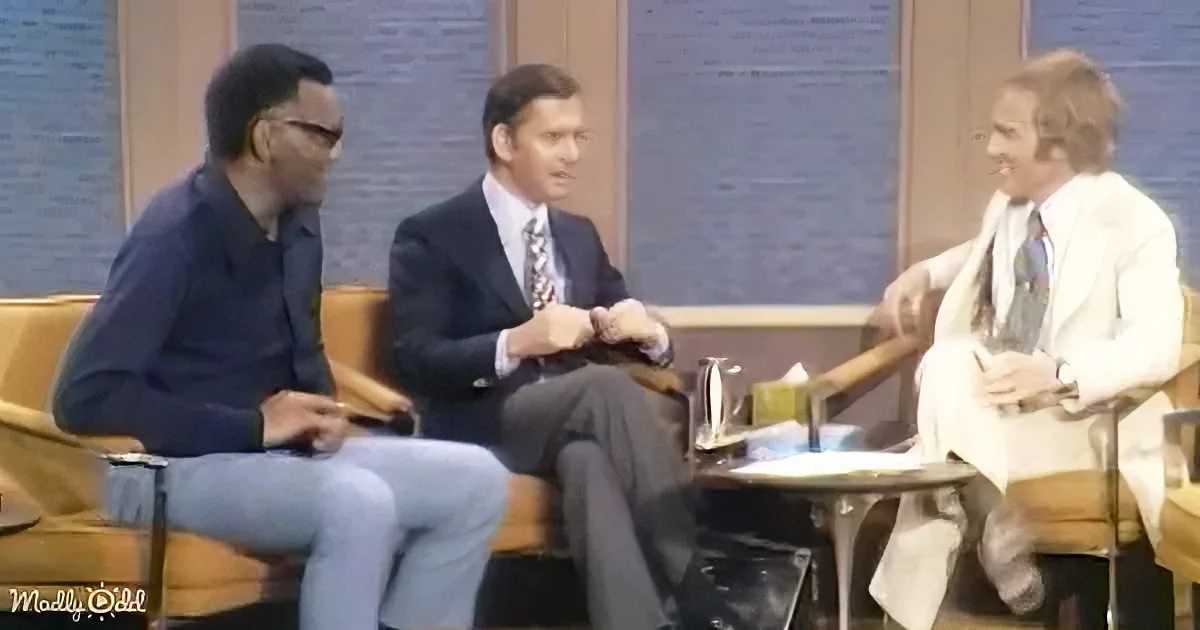 Three men in suits sit on a talk show set, engaged in conversation.