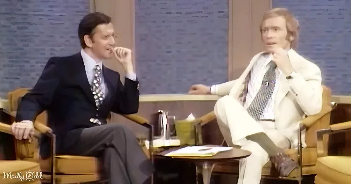 Two men in suits sit on chairs, one in black, the other in white, engaged in conversation.
