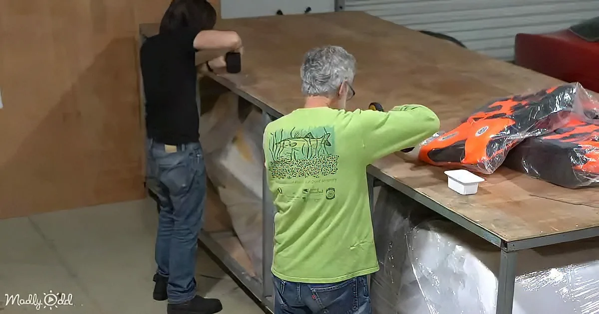 A man in a green shirt works on a wooden structure in a workshop, with another person nearby.