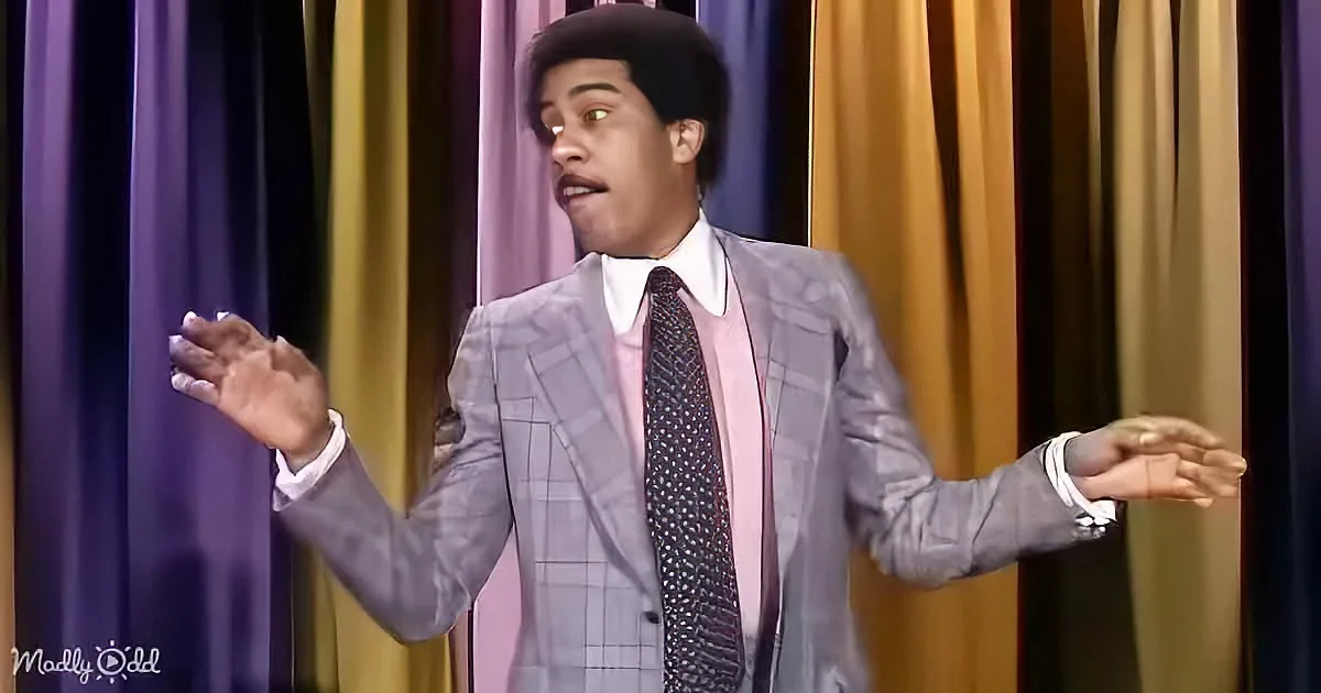 A man in a checkered suit and tie stands with arms outstretched, looking surprised, against a colorful curtain backdrop.
