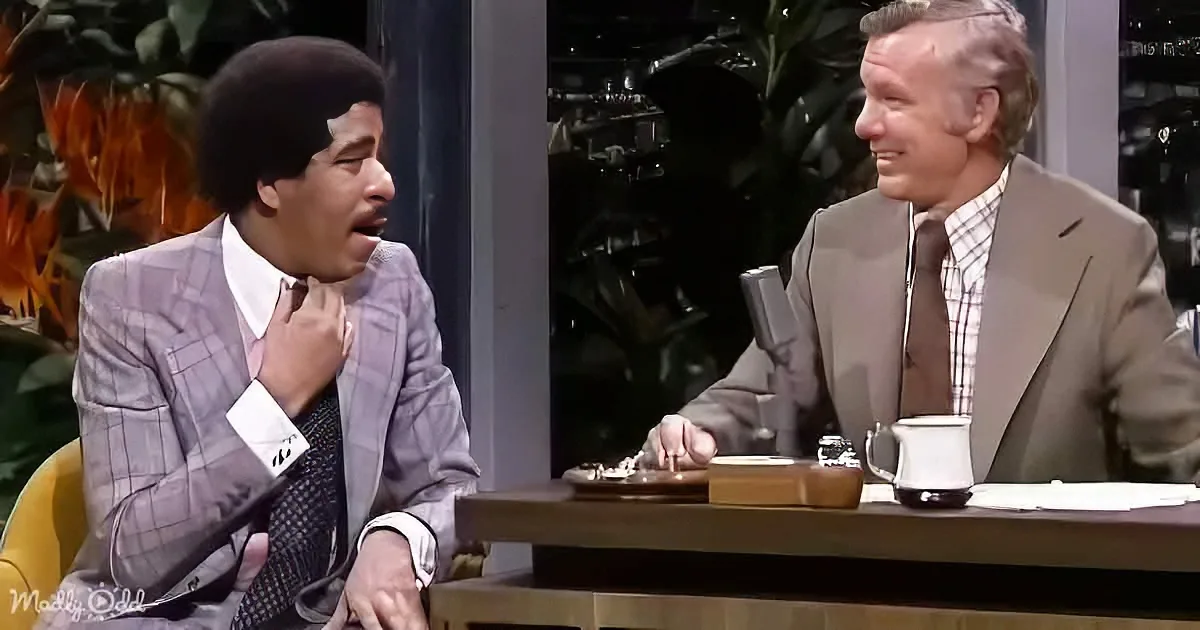 A man in a plaid suit adjusts his tie while another man in a suit looks on during a talk show.
