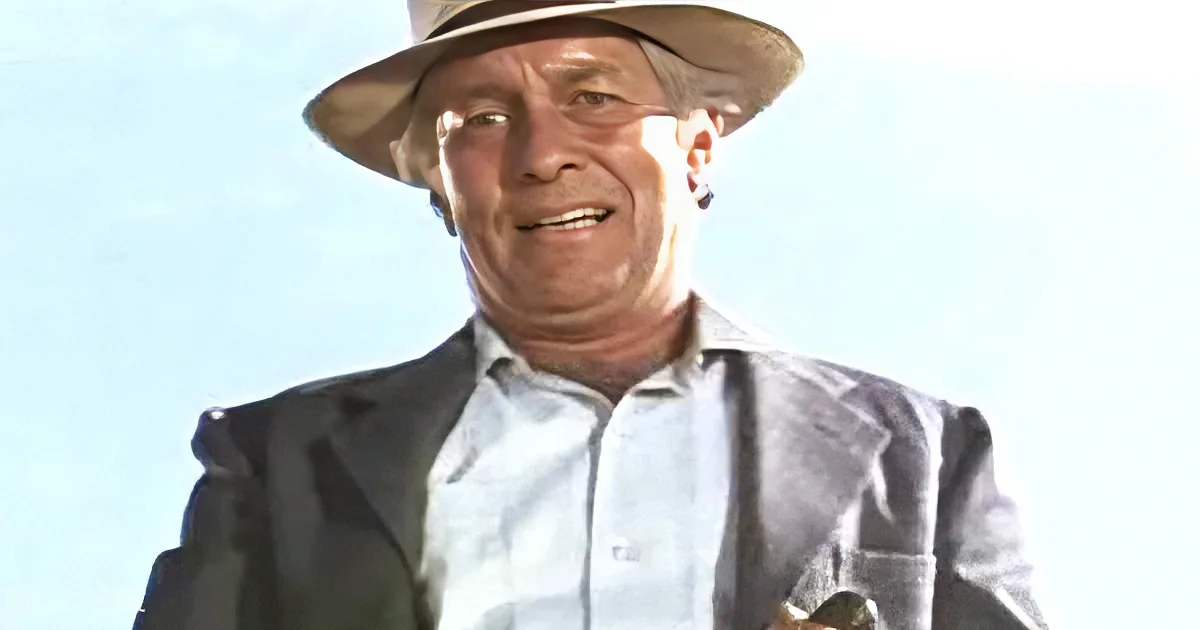 A man in a hat and suit smiles, wearing a light blue shirt and a dark jacket.