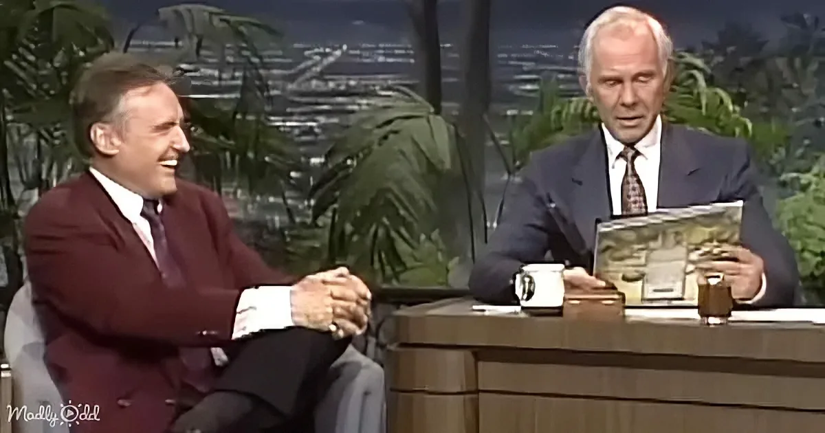 A man in a maroon suit laughs while seated next to a man in a suit holding a book on a talk show set.