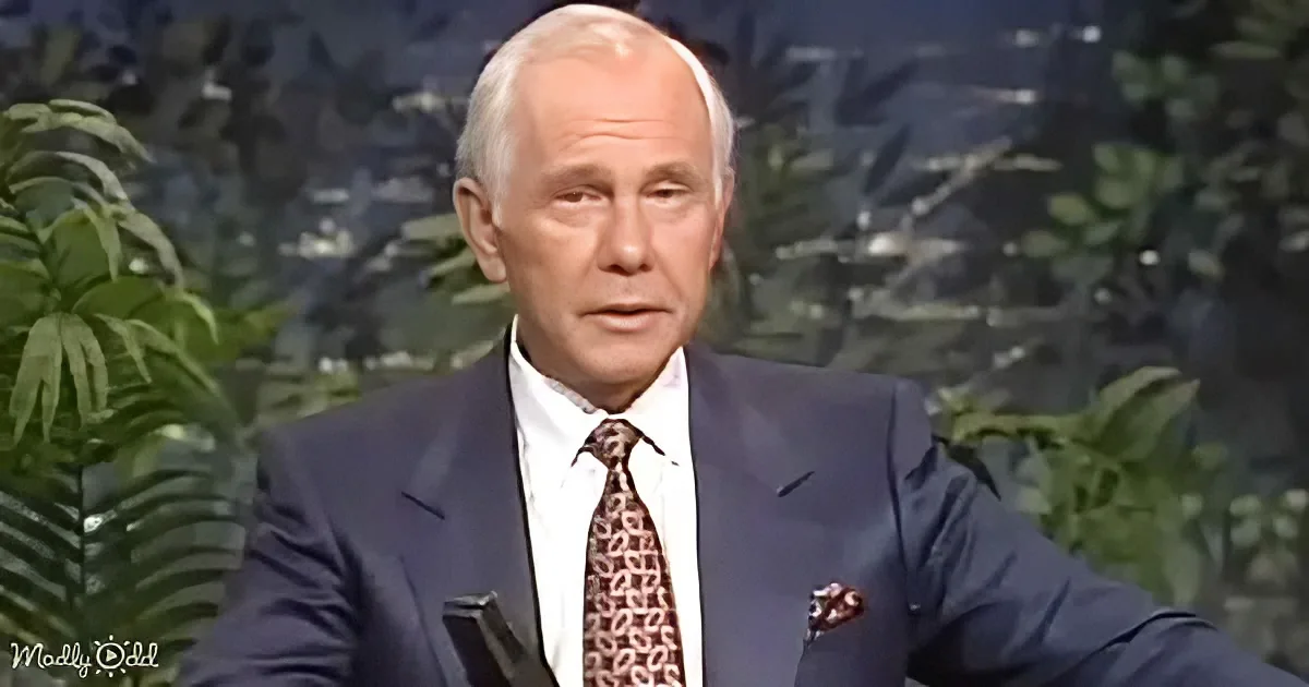 A man in a suit and tie sits against a backdrop of greenery, appearing to speak.