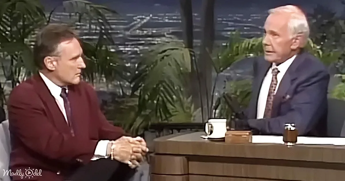 Two men in suits sit across from each other on a talk show set with a cityscape backdrop.