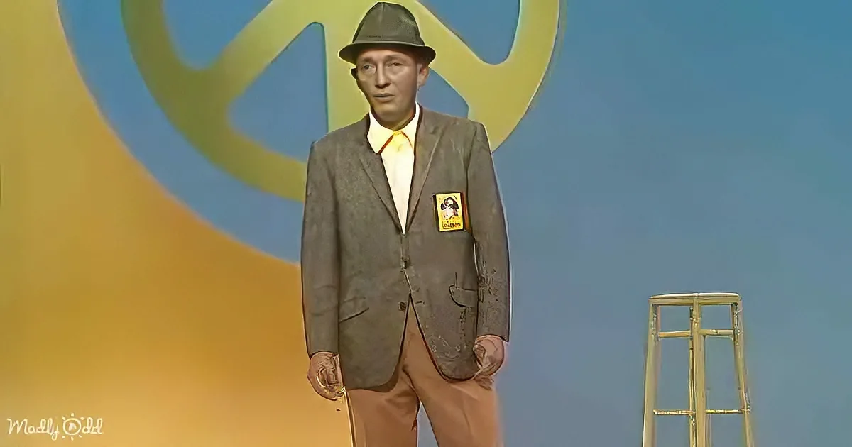 A man in a suit and hat stands on stage, wearing a name tag, with a colorful backdrop and a stool in the background.