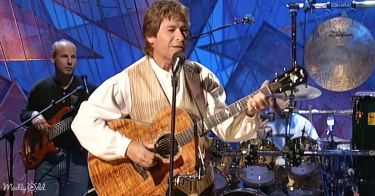 A man in a striped shirt plays an acoustic guitar on stage with a microphone and other musicians.