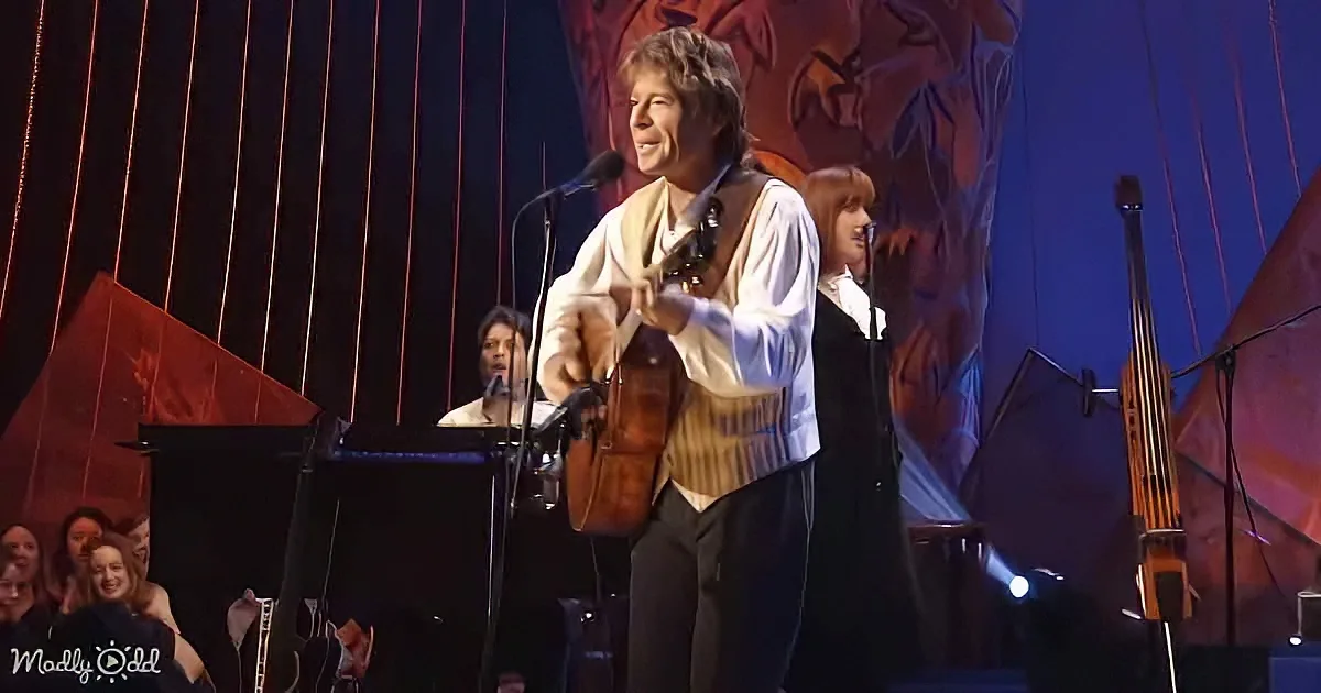A man in a white shirt and vest plays a guitar on stage with a microphone, surrounded by musicians and a red backdrop.