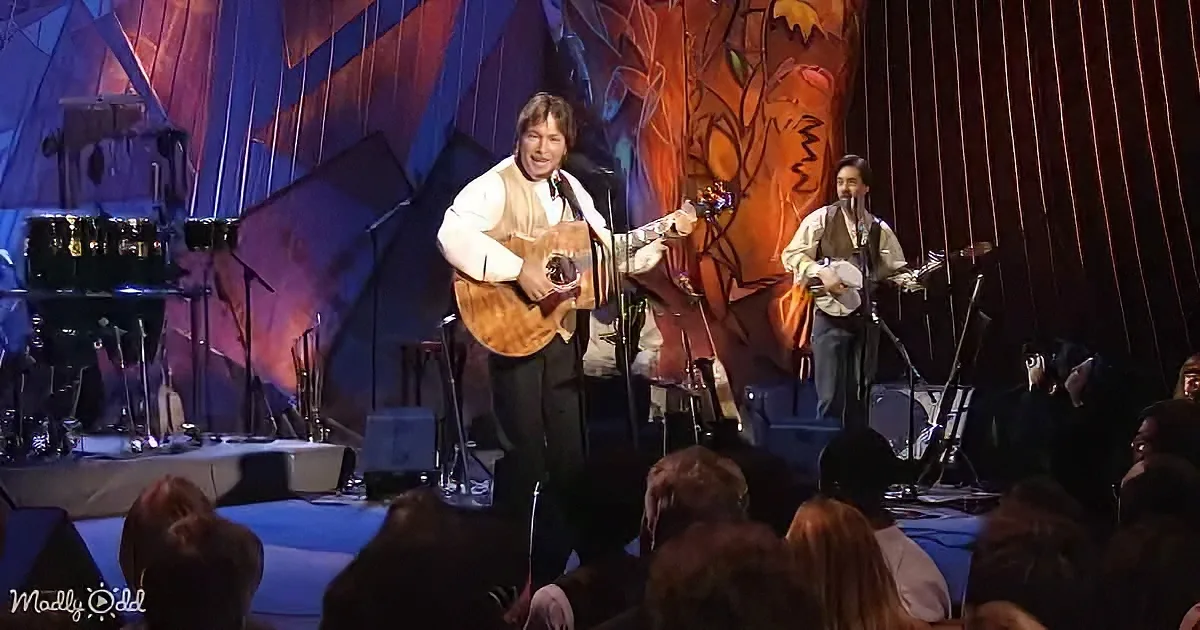 Two musicians perform on stage with acoustic guitars, surrounded by an audience and colorful backdrop.