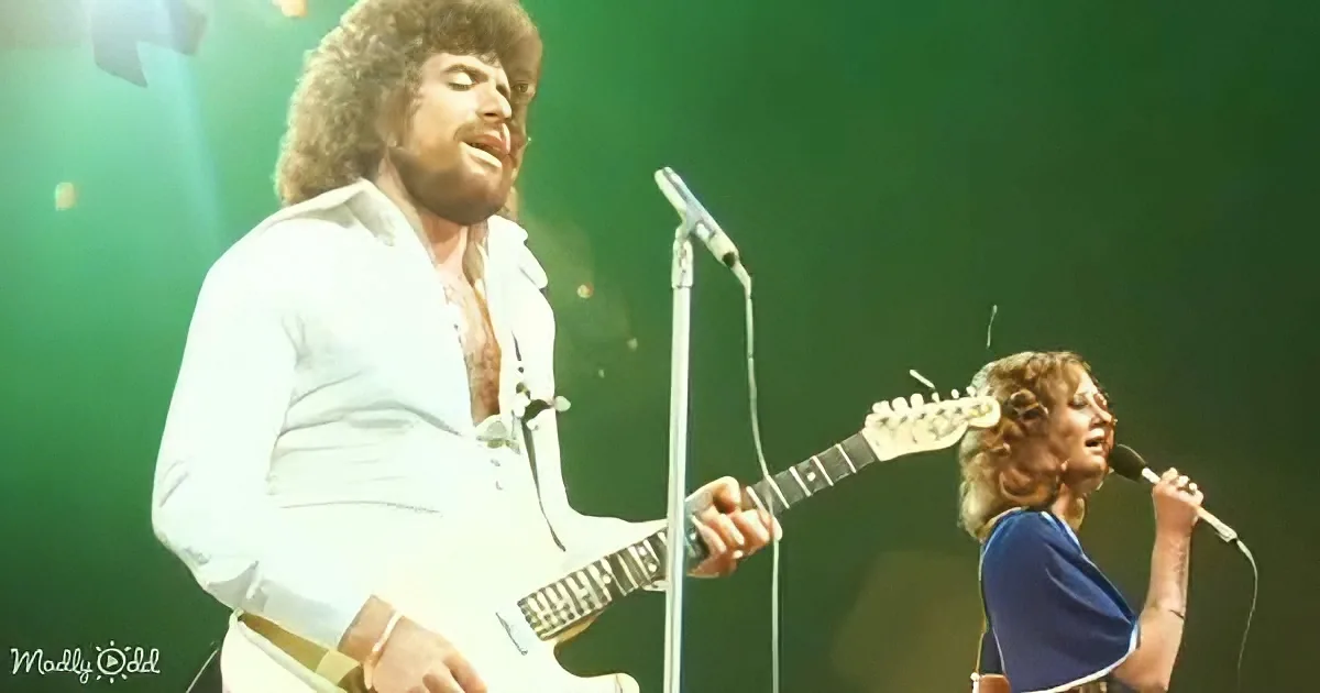 A man with curly hair plays guitar while a woman sings into a microphone on stage.
