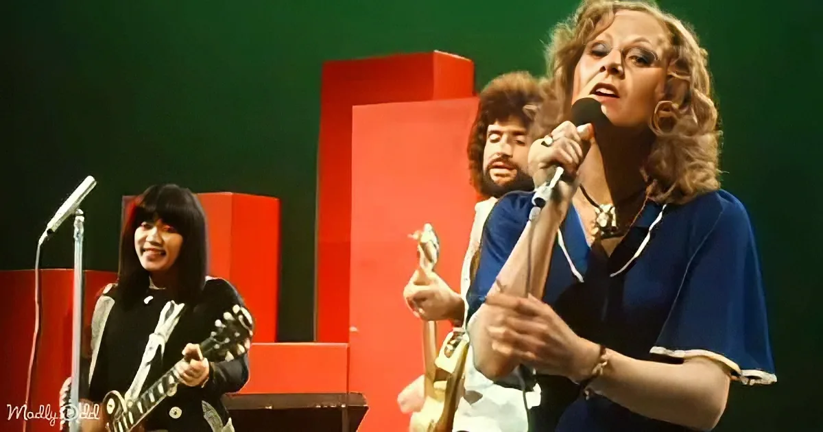 A woman in a blue outfit sings into a microphone on stage with two musicians behind her.