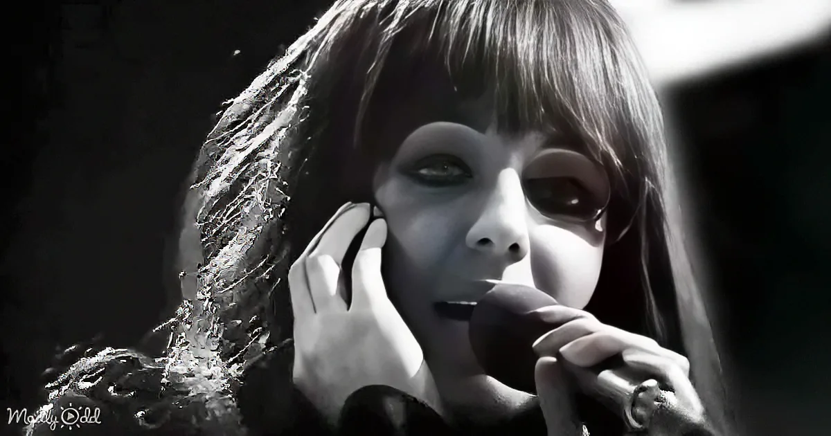 A person with long hair and pale makeup holds a microphone, creating a dramatic and eerie atmosphere.