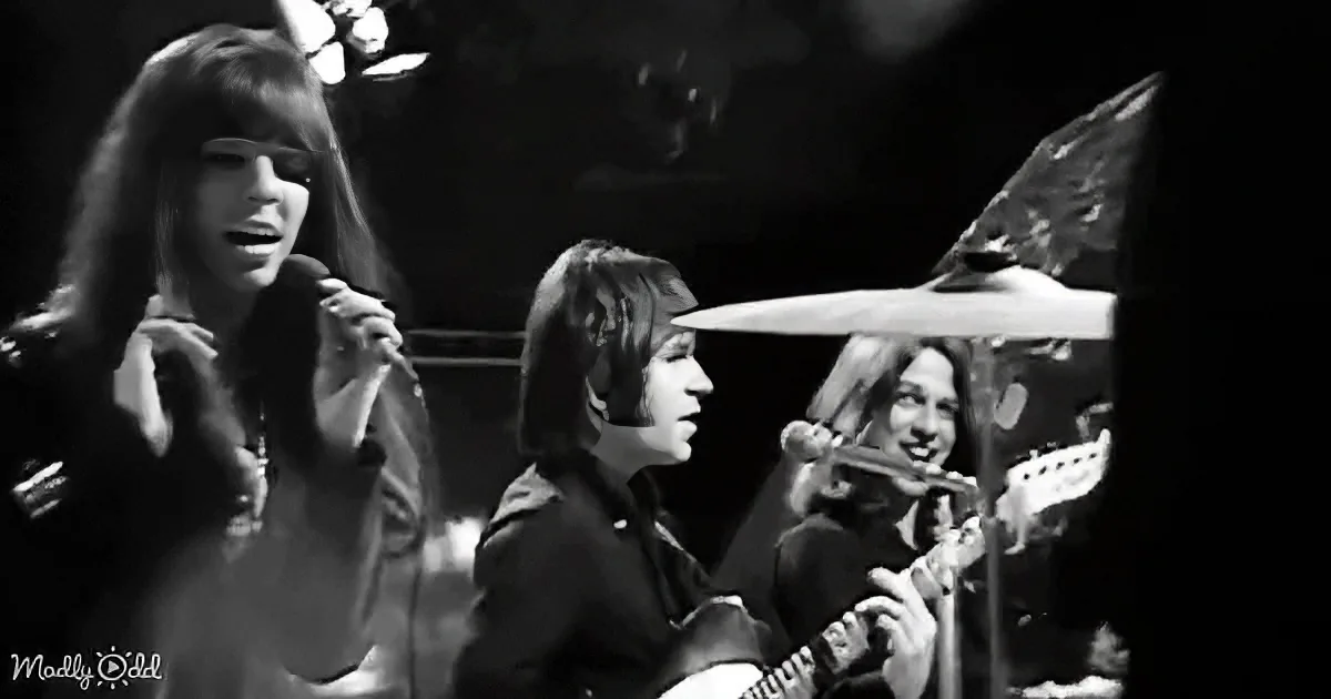 A woman sings into a microphone while a guitarist and drummer play in the background.