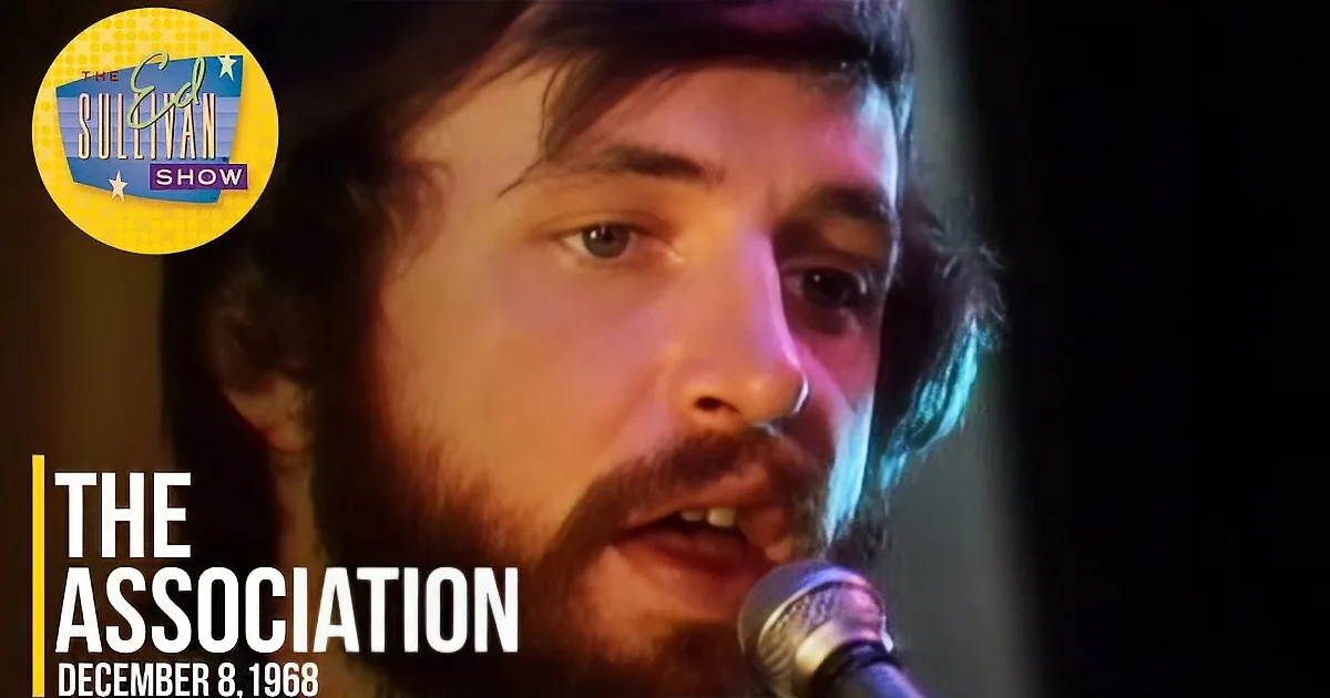 A man with a beard and dark hair sings into a microphone, illuminated by warm lighting.