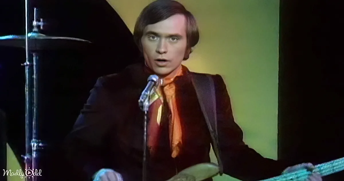 A man in a black suit and orange tie sings into a microphone, holding a guitar, with a drum set in the background.