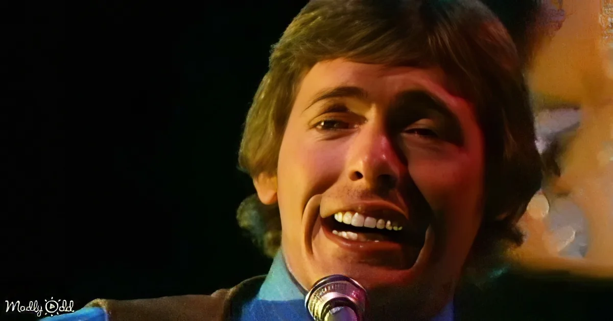 A man with shoulder-length hair sings into a microphone, wearing a blue shirt, against a dark background.