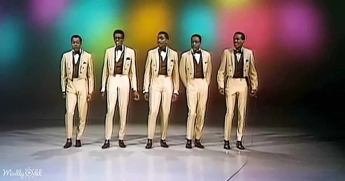 Five men in cream suits stand in a line against a colorful, blurred background.