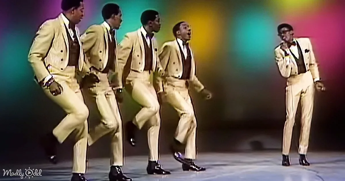 A group of five men in matching beige suits perform a synchronized dance routine on stage.