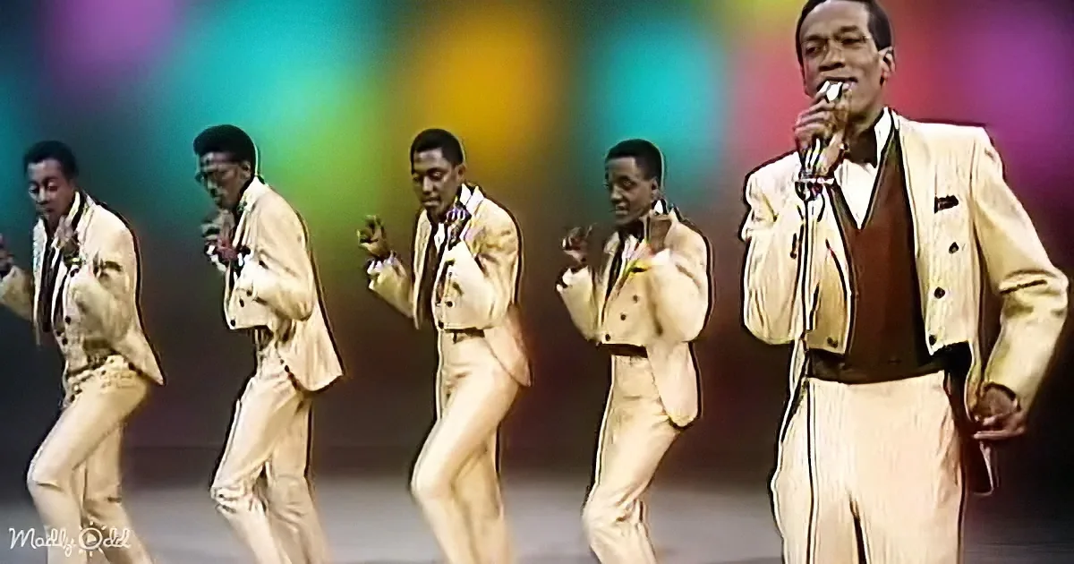 A man in a cream suit sings into a microphone, surrounded by four dancers in similar attire.