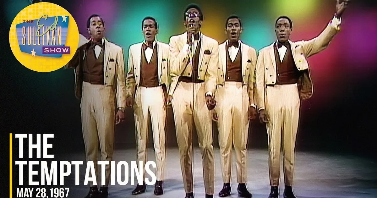 A man in a white suit sings into a microphone, surrounded by four other men in similar attire.