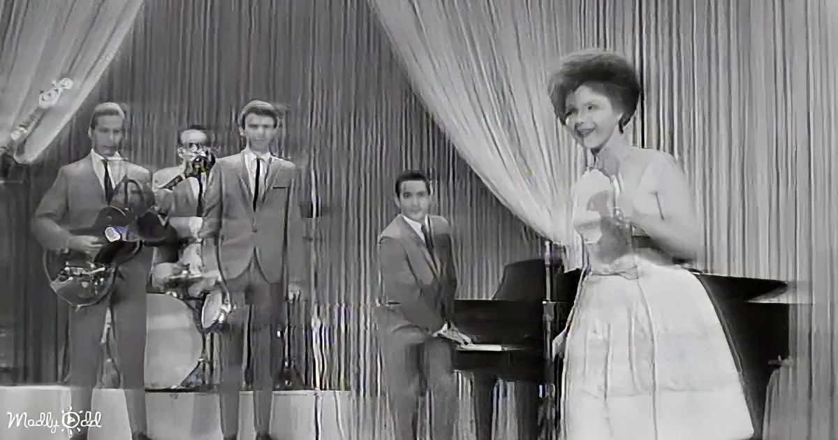 A woman in a white dress sings into a microphone, accompanied by a band in suits on stage.