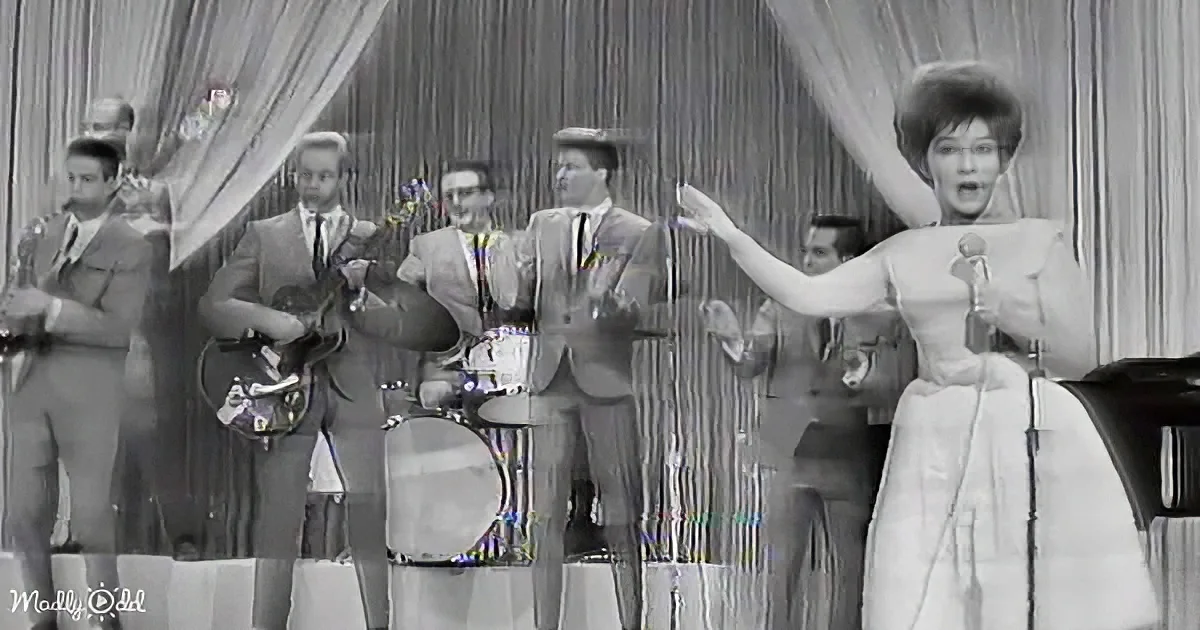 A woman in a white dress sings into a microphone, gesturing dramatically, with a band behind her.