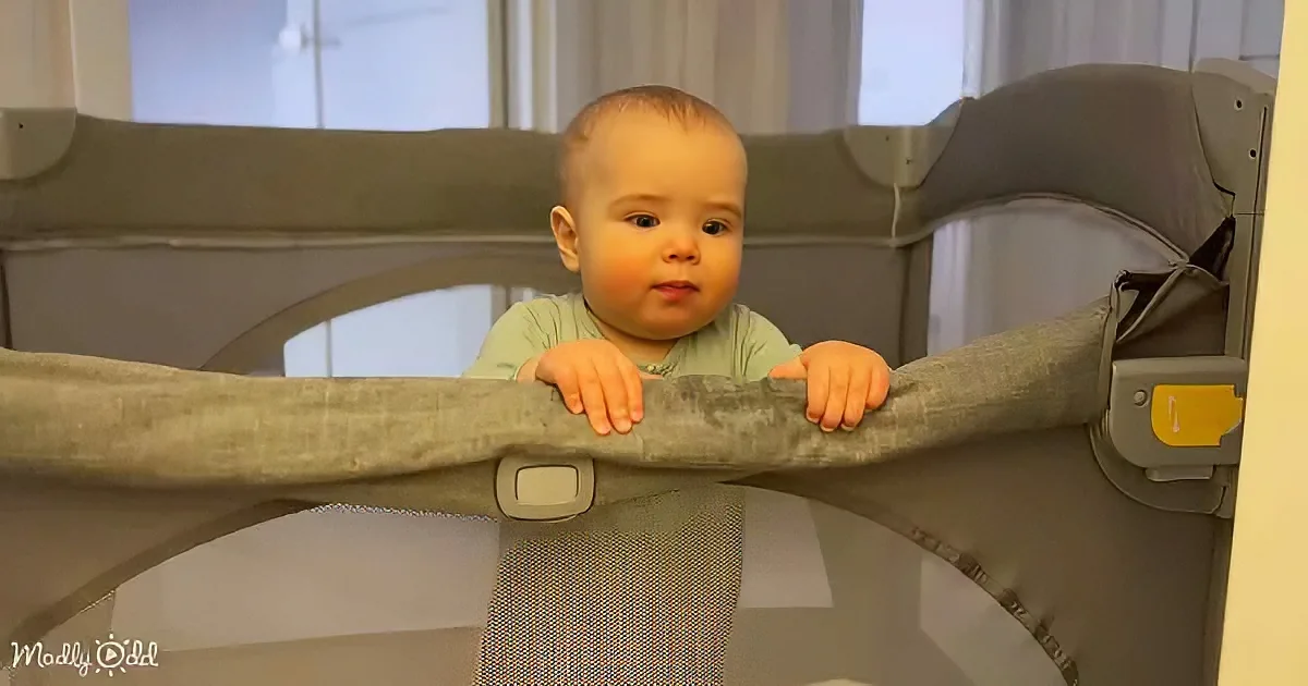 A baby in a green shirt stands in a gray playpen, holding onto the top rail.