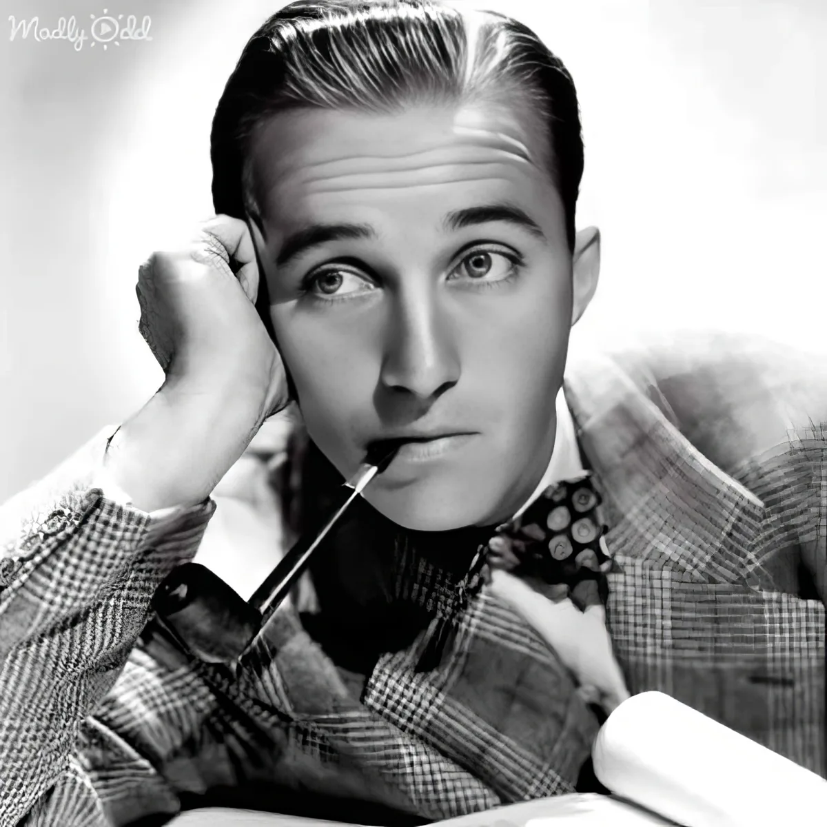 A man in a checkered suit and bow tie holds a cigarette, hand on his head, gazing upward.