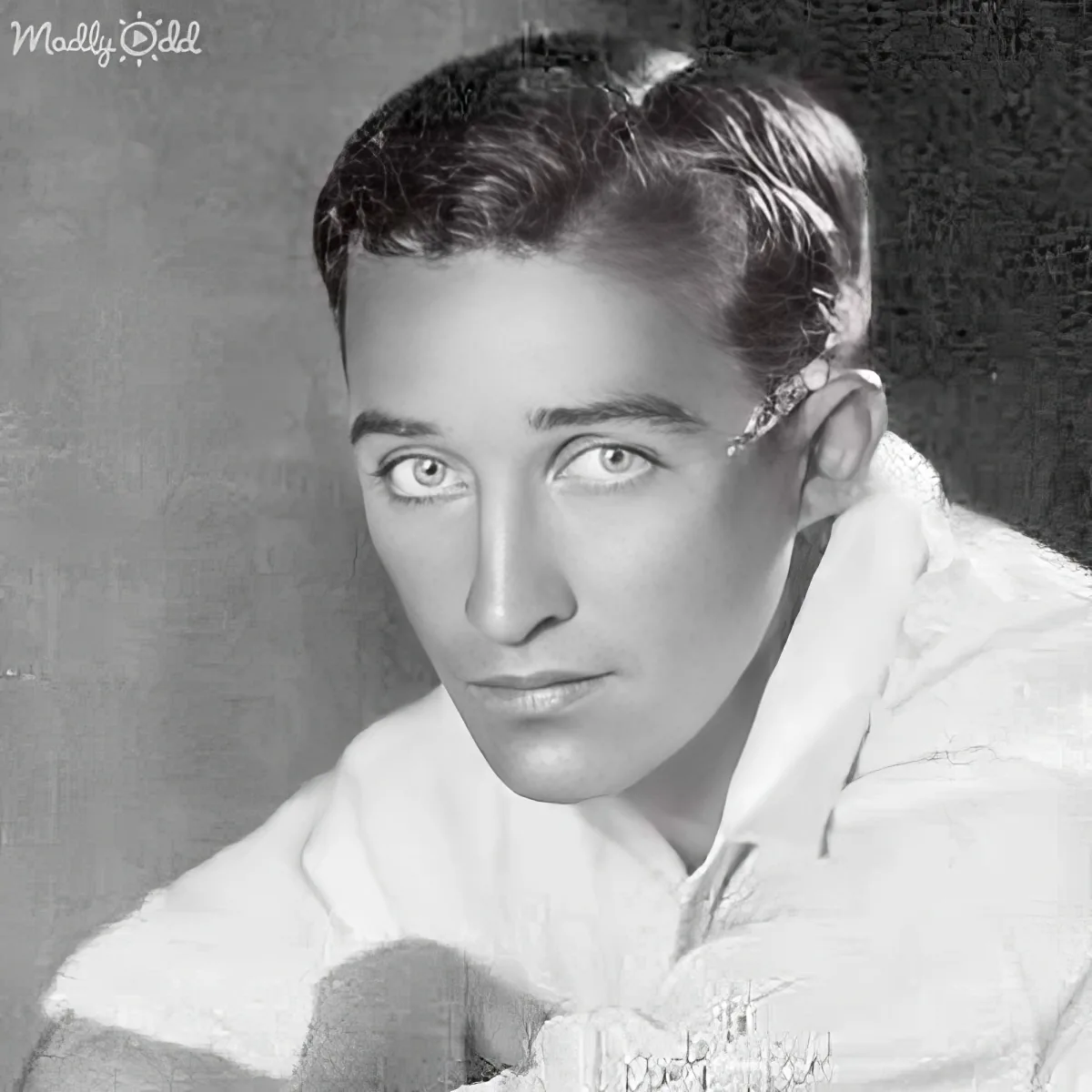 A man with slicked-back hair and a white shirt gazes directly at the camera in a black-and-white portrait.