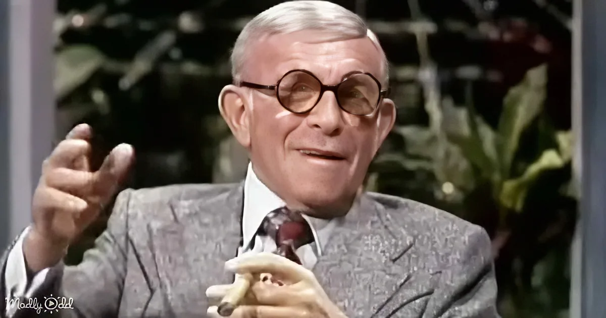An older man in a suit and tie, wearing glasses, holds a banana and gestures with his right hand.