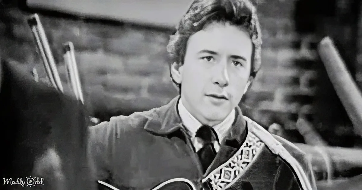 A man with curly hair, wearing a jacket and tie, holds a guitar, looking off to the side.