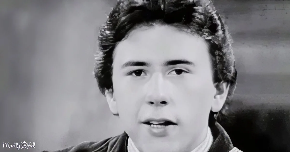 A man with short, curly hair and a serious expression is shown in a black-and-white close-up.