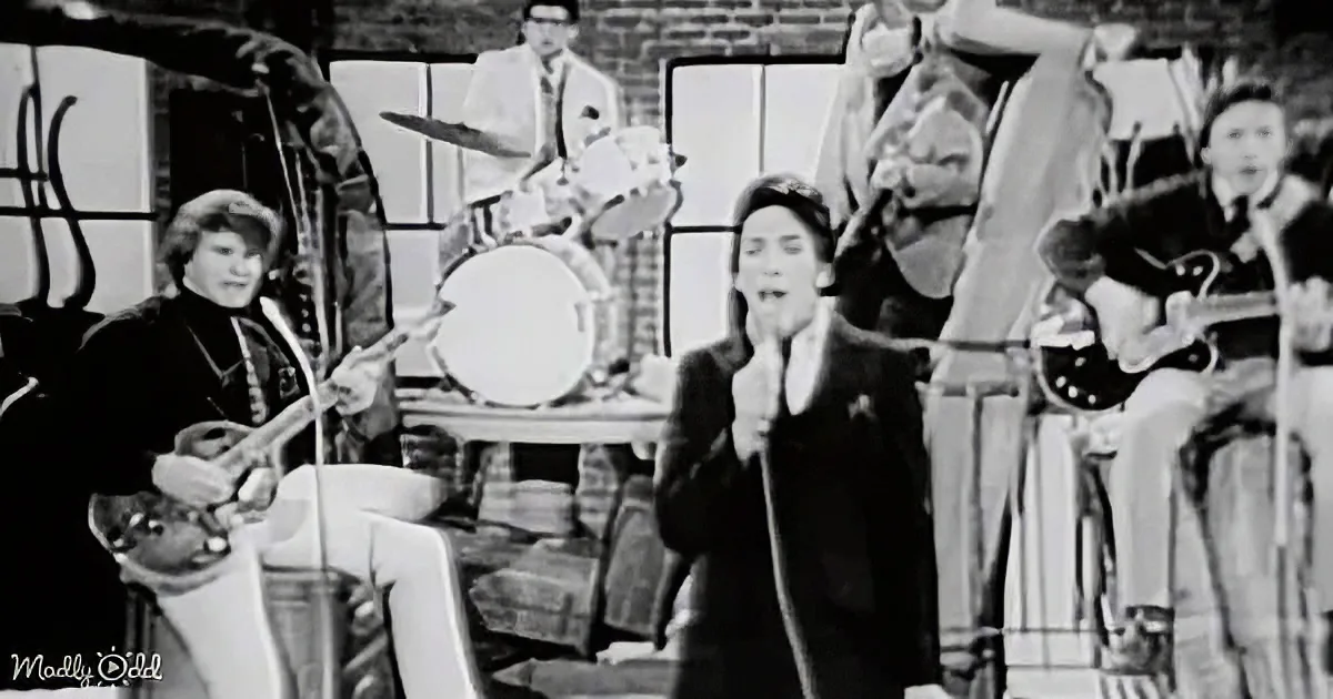 A man in a suit sings into a microphone while a band plays behind him in a vintage setting.