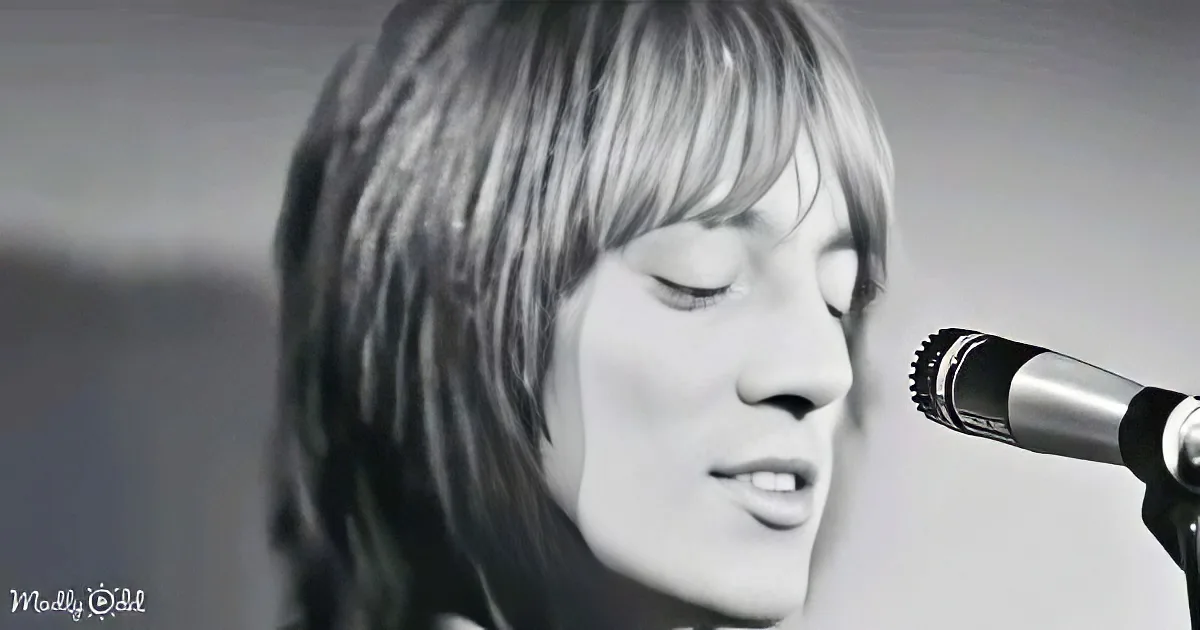 A person with shoulder-length hair sings into a microphone, eyes closed, in a close-up shot.