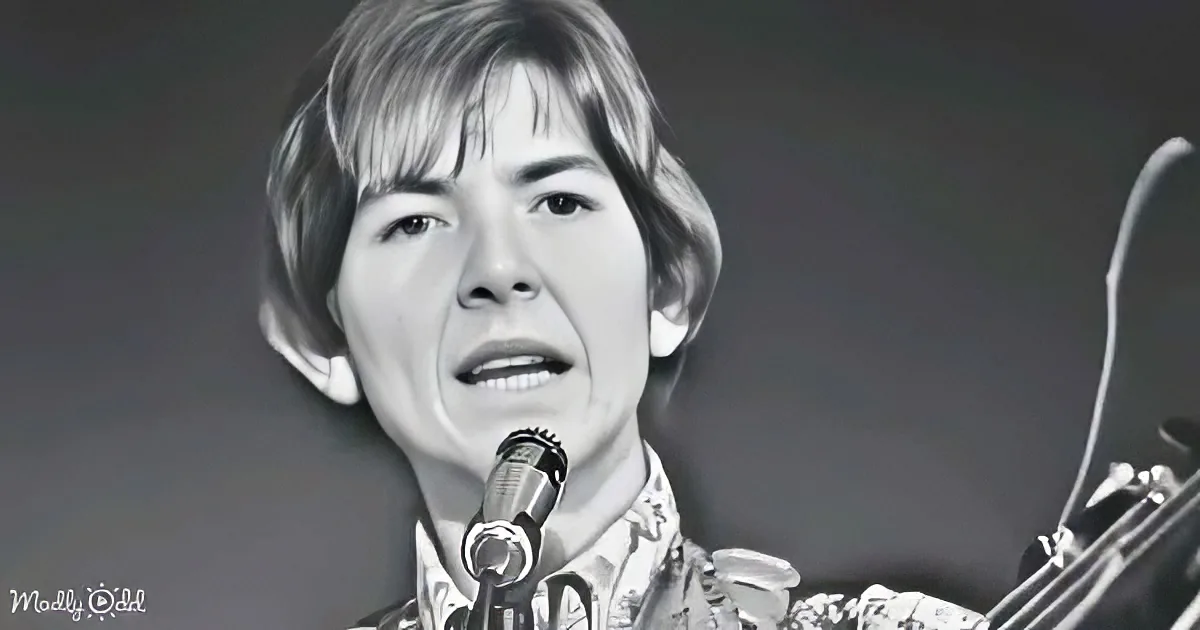 A person singing into a microphone, wearing a patterned shirt, with a blurred background.
