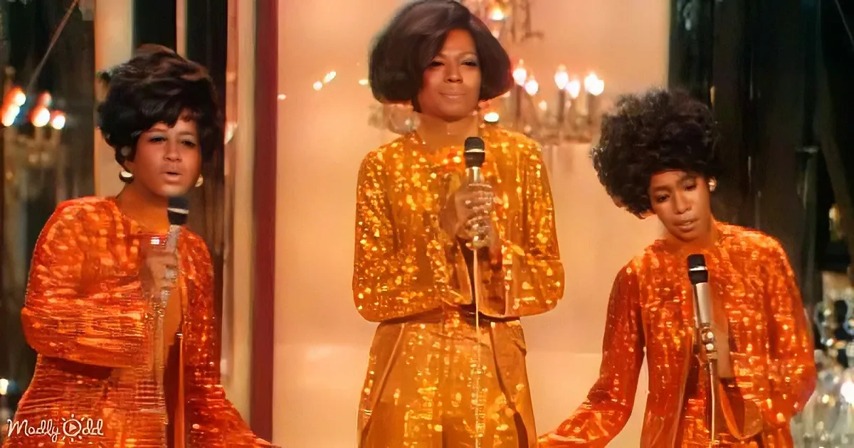Three women in shimmering orange outfits sing into microphones on a stage with chandeliers.