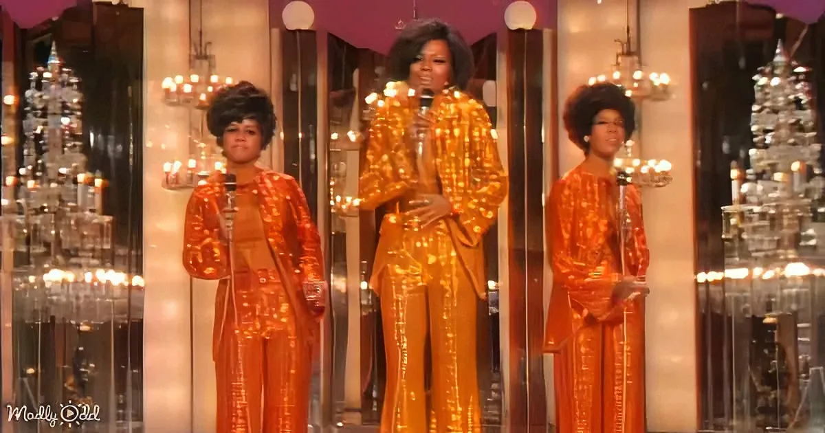 A woman in a golden sequined outfit sings on stage, flanked by two backup dancers in matching orange outfits.