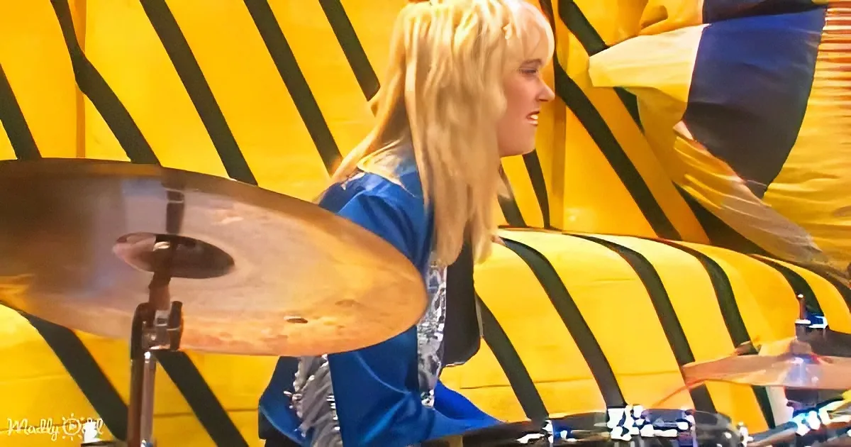 A person with long blonde hair in a blue outfit plays drums in front of a yellow and black striped background.