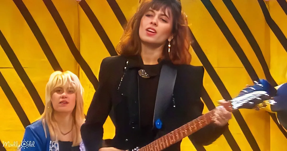 A woman in a black outfit plays guitar, singing, with another person in a blue shirt behind her.