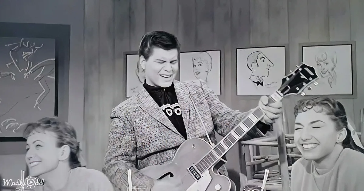 A man in a patterned suit plays a guitar, surrounded by smiling women and caricature drawings on the wall.