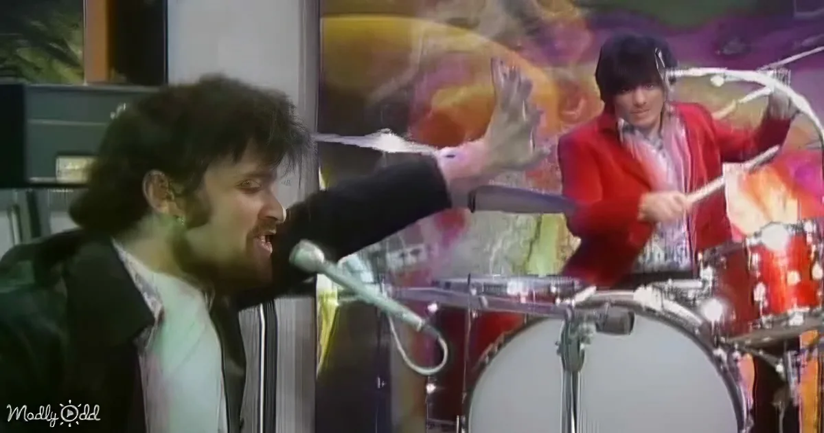 A man in a green jacket sings passionately into a microphone while a drummer in a red jacket plays in the background.