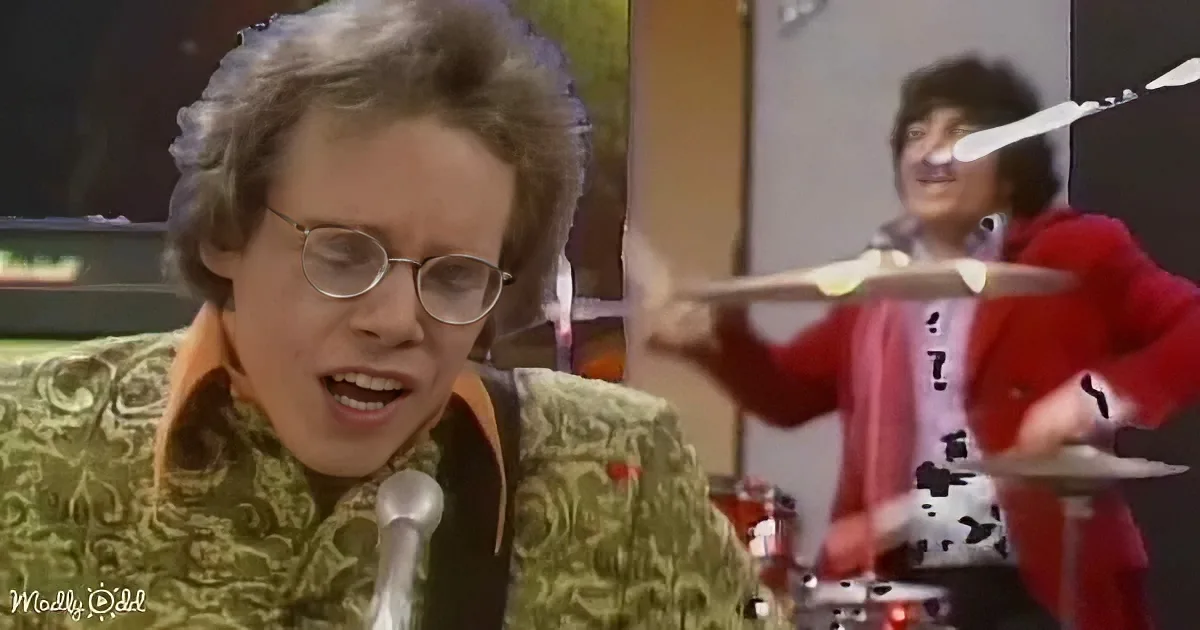 A man in a green patterned shirt sings into a microphone while another person plays drums in the background.