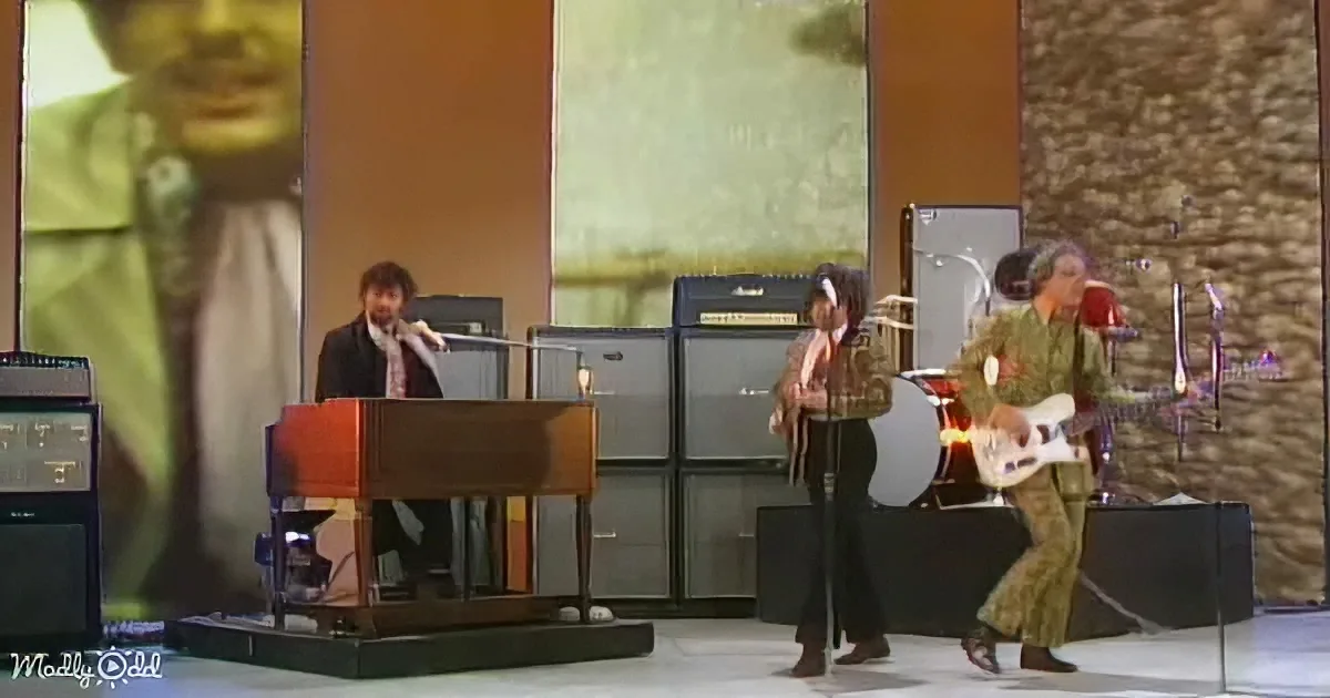 A man plays piano on stage with a band, large screen behind him.