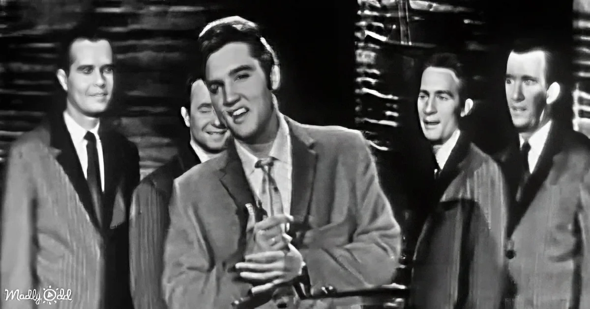 Elvis Presley, center, in a suit, smiling, surrounded by four men in suits, black-and-white.
