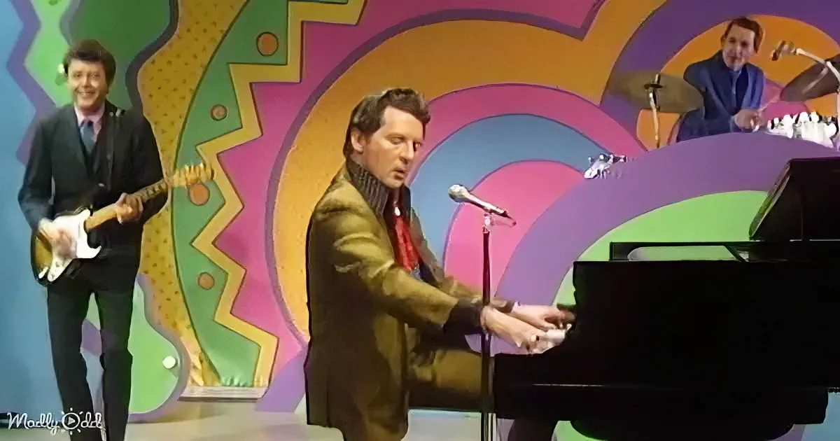 A man in a brown suit plays piano on stage with a colorful background and other musicians.